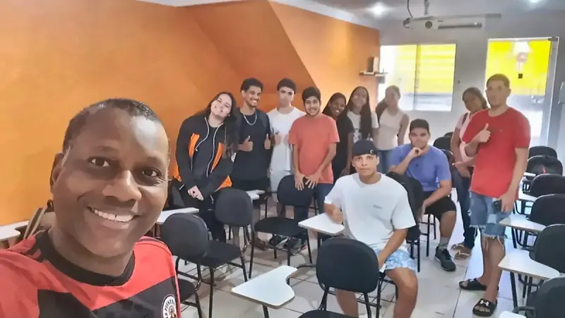 Selfie sorridente com a turma do Foco ao fundo, na sala de parede laranja